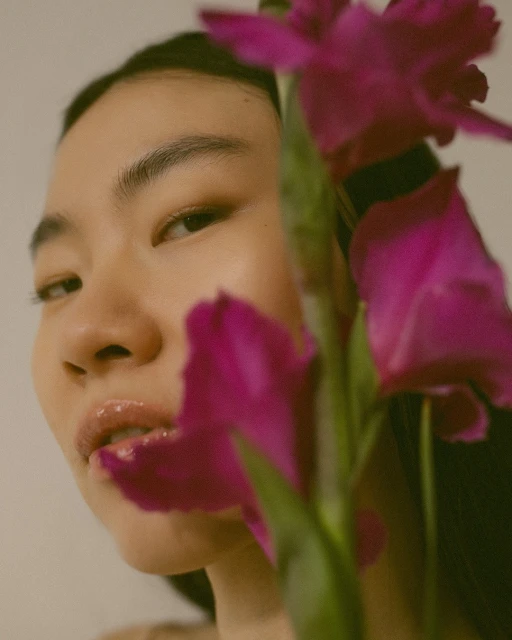 Close-up portrait of a young Asian woman with glossy lips partially obscured by magenta-colored gladiolus flowers against a light backdrop.
