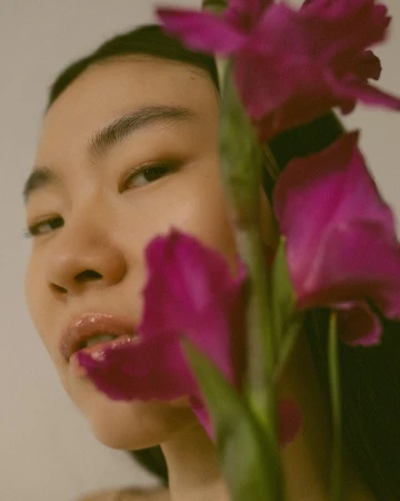 Close-up portrait of a young Asian woman with glossy lips partially obscured by magenta-colored gladiolus flowers against a light backdrop.