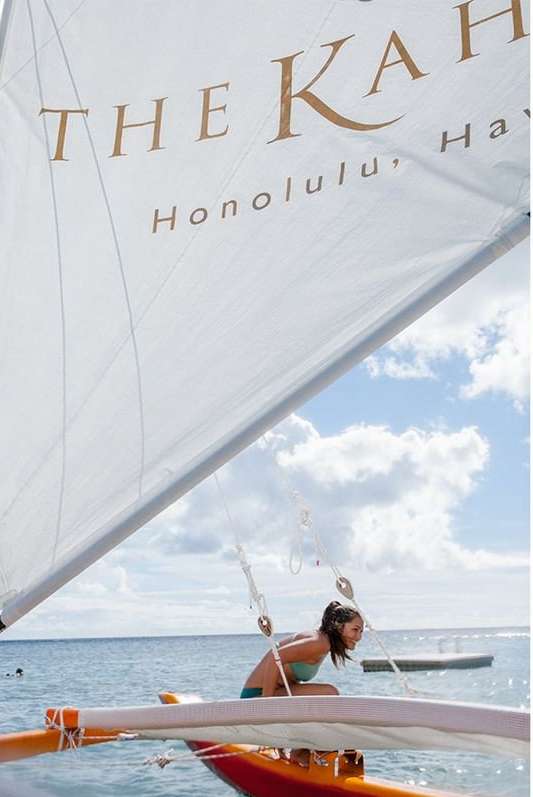 A woman sailing a catamaran on the ocean near Honolulu, Hawaii, with a sail printed with "The Kahala, Honolulu, Hawaii".