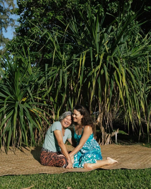 Two women, one older with short gray hair and one younger with long dark hair, sit together on a woven mat in a green garden.