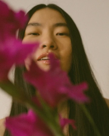 Close-up portrait of a woman partially obscured by vibrant magenta flowers in the foreground, photographed by Daeja Fallas.
