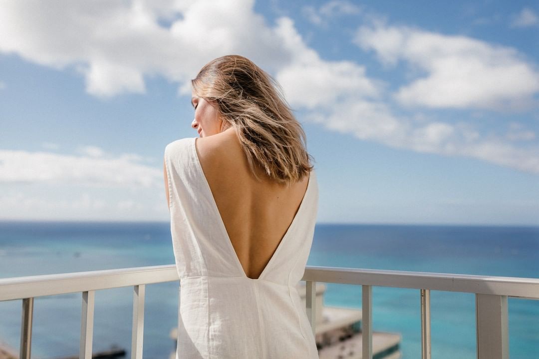 A woman in a white dress stands on a balcony overlooking the ocean. The sky is blue with clouds. The woman's back is exposed.