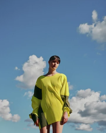 A woman wearing a vibrant yellow and dark green patterned top posing against a bright blue sky with white clouds.