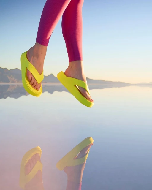 Woman in pink leggings jumps over reflective water, wearing bright yellow flip flops; mountains in background, sunny day.