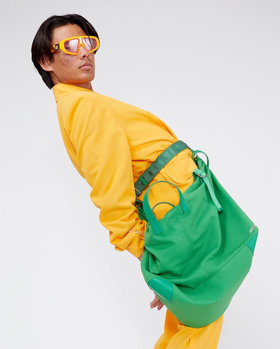 Adult male posing in front of a white backdrop, wearing yellow clothing and orange sunglasses with a green shoulder bag.