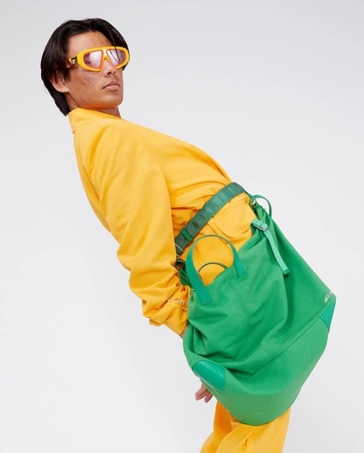 Adult male posing in front of a white backdrop, wearing yellow clothing and orange sunglasses with a green shoulder bag.