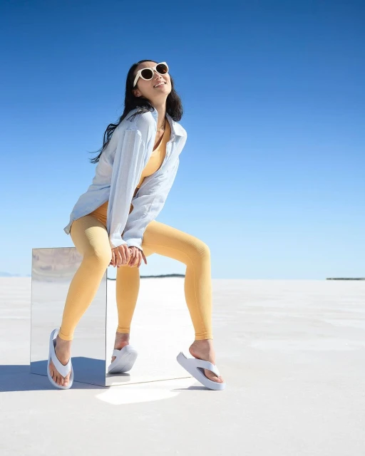 A woman sits on a reflective block in a desert landscape, wearing sunglasses, a yellow top and leggings, and slides, under a blue sky.