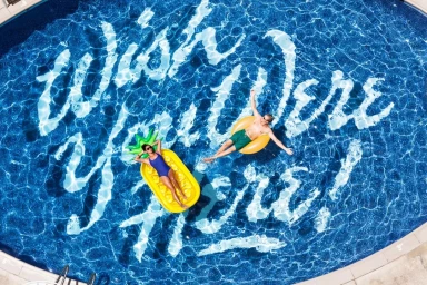 Aerial view of a swimming pool with large white typography reading Wish You Were Here across the bottom, with two people floating on inflatables.