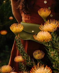 Close-up of a person in a rust-orange dress wearing an olive green leather fanny pack amidst bright orange pincushion flowers