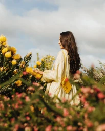 Woman in pale dress with a yellow crossbody bag touching yellow flowers in a garden photographed by Daeja Fallas.