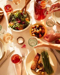 Overhead view of a sunlit dining table with salmon, asparagus, salad, wine glasses, and hands serving food.