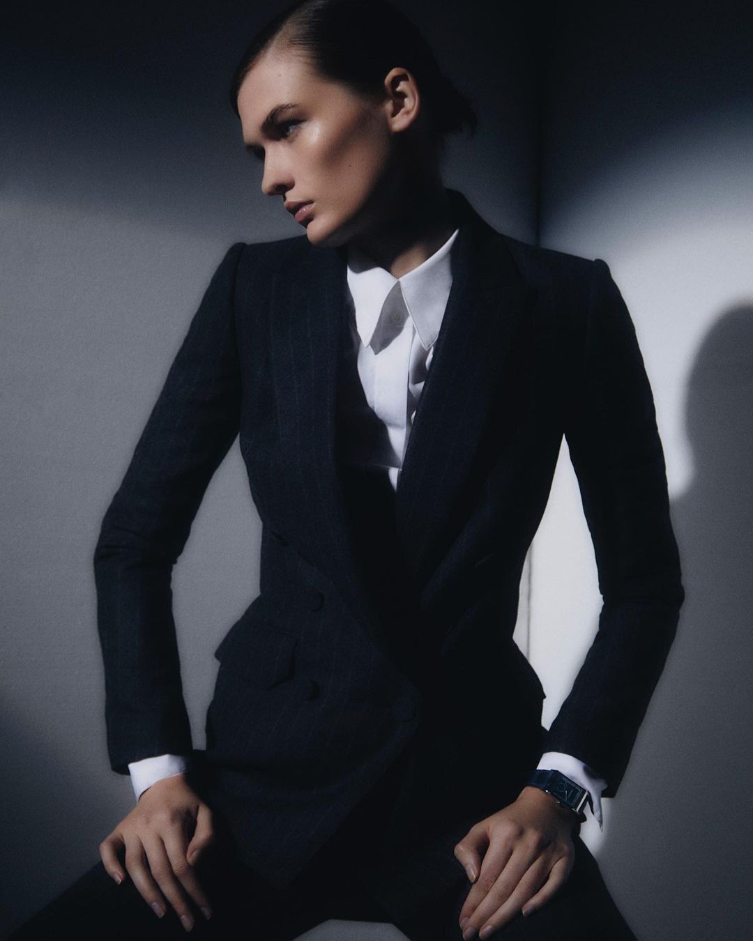 Model wearing a dark blazer over a white collared shirt seated against a grey studio wall with dramatic low-key lighting