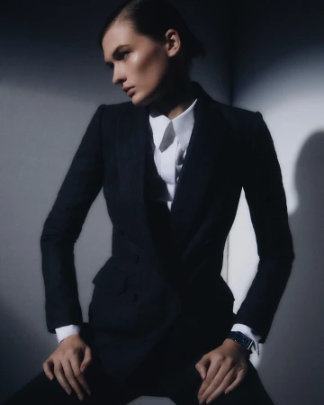 Model wearing a dark blazer over a white collared shirt seated against a grey studio wall with dramatic low-key lighting