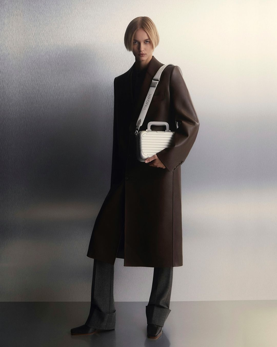 Fashion shot of a woman in a brown coat holding a silver colored case with gray pants and black boots in a neutral studio environment.