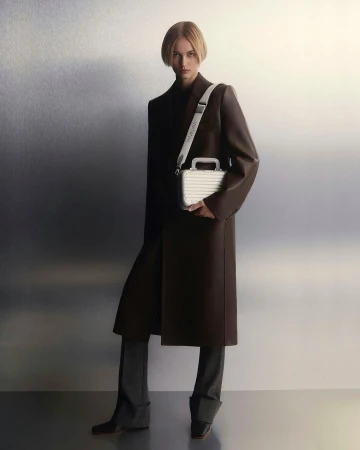 Fashion shot of a woman in a brown coat holding a silver colored case with gray pants and black boots in a neutral studio environment.