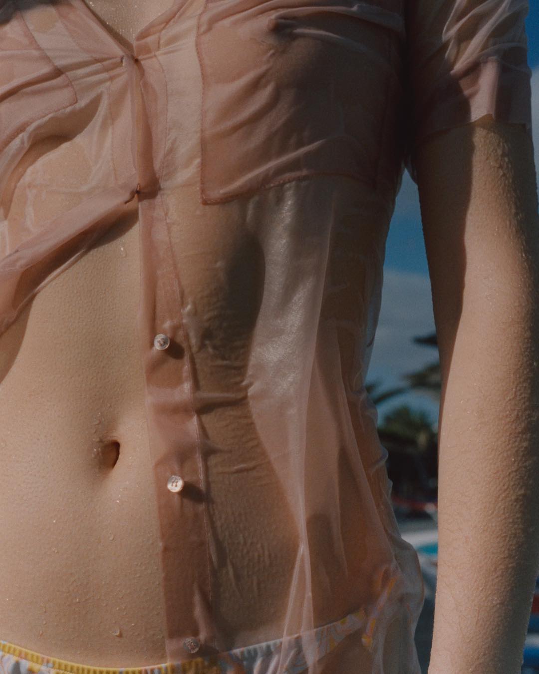 Close-up of a female torso wearing a wet, sheer peach blouse in an outdoor setting near water and palm trees during daytime.
