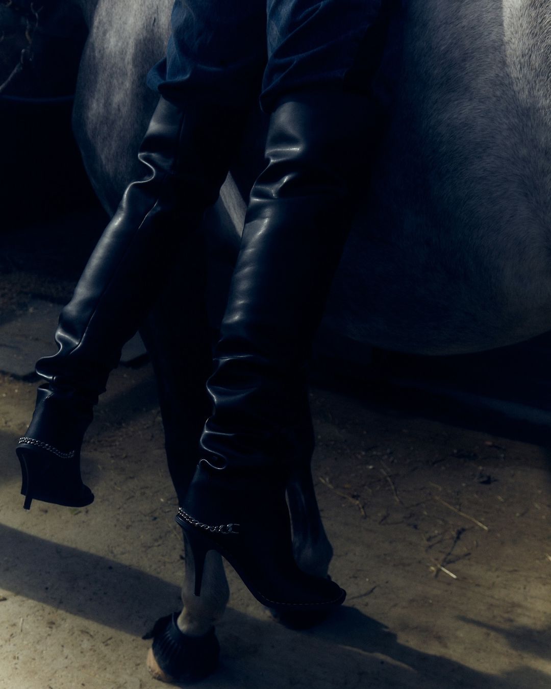 Woman wearing black heeled boots while sitting astride a grey horse in an indoor stable setting. Fashion and equine lifestyle.