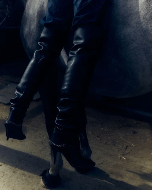 Woman wearing black heeled boots while sitting astride a grey horse in an indoor stable setting. Fashion and equine lifestyle.
