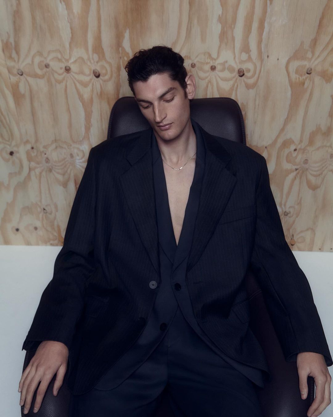Man wearing a dark oversized blazer and matching trousers seated in a black leather chair against a plywood wall