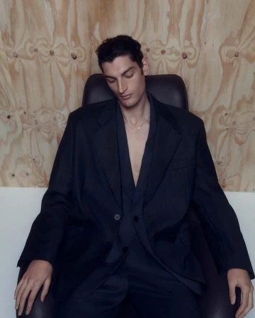 Man wearing a dark oversized blazer and matching trousers seated in a black leather chair against a plywood wall