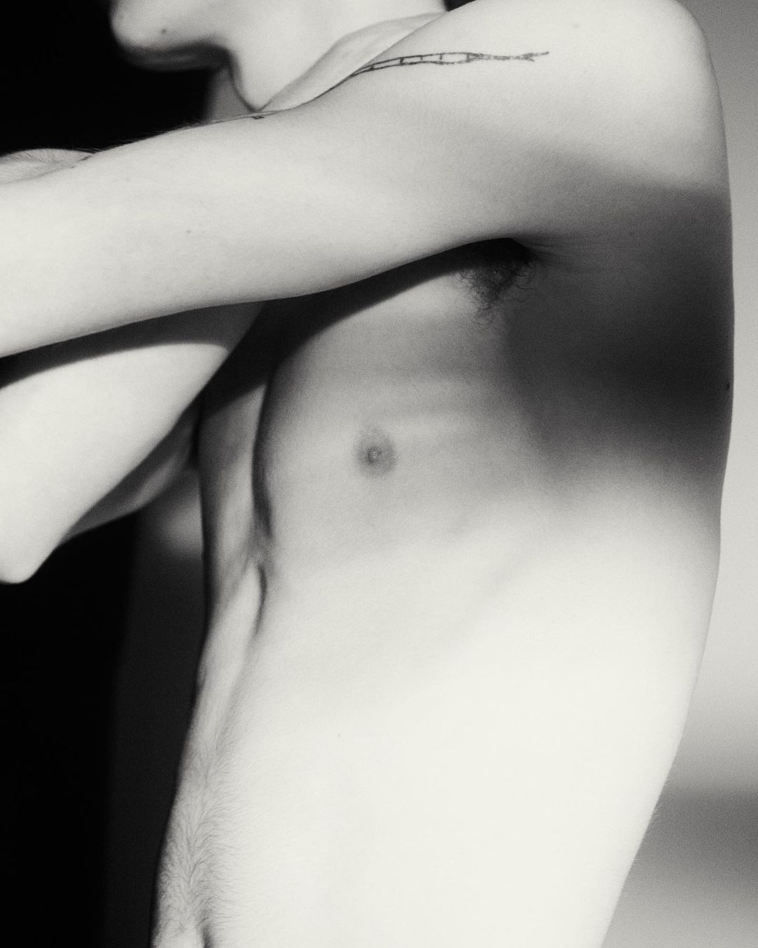 Black and white portrait of shirtless adult male torso and arm with linear tattoo, set against a gradated neutral background.