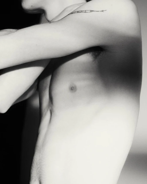 Black and white portrait of shirtless adult male torso and arm with linear tattoo, set against a gradated neutral background.