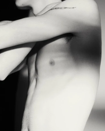 Black and white portrait of shirtless adult male torso and arm with linear tattoo, set against a gradated neutral background.