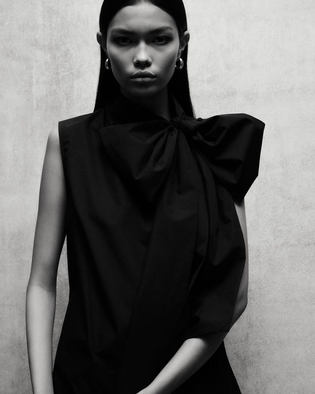 Portrait of a model wearing a black sleeveless top with a large bow detail and hoop earrings against a textured backdrop