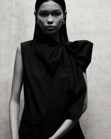 Portrait of a model wearing a black sleeveless top with a large bow detail and hoop earrings against a textured backdrop