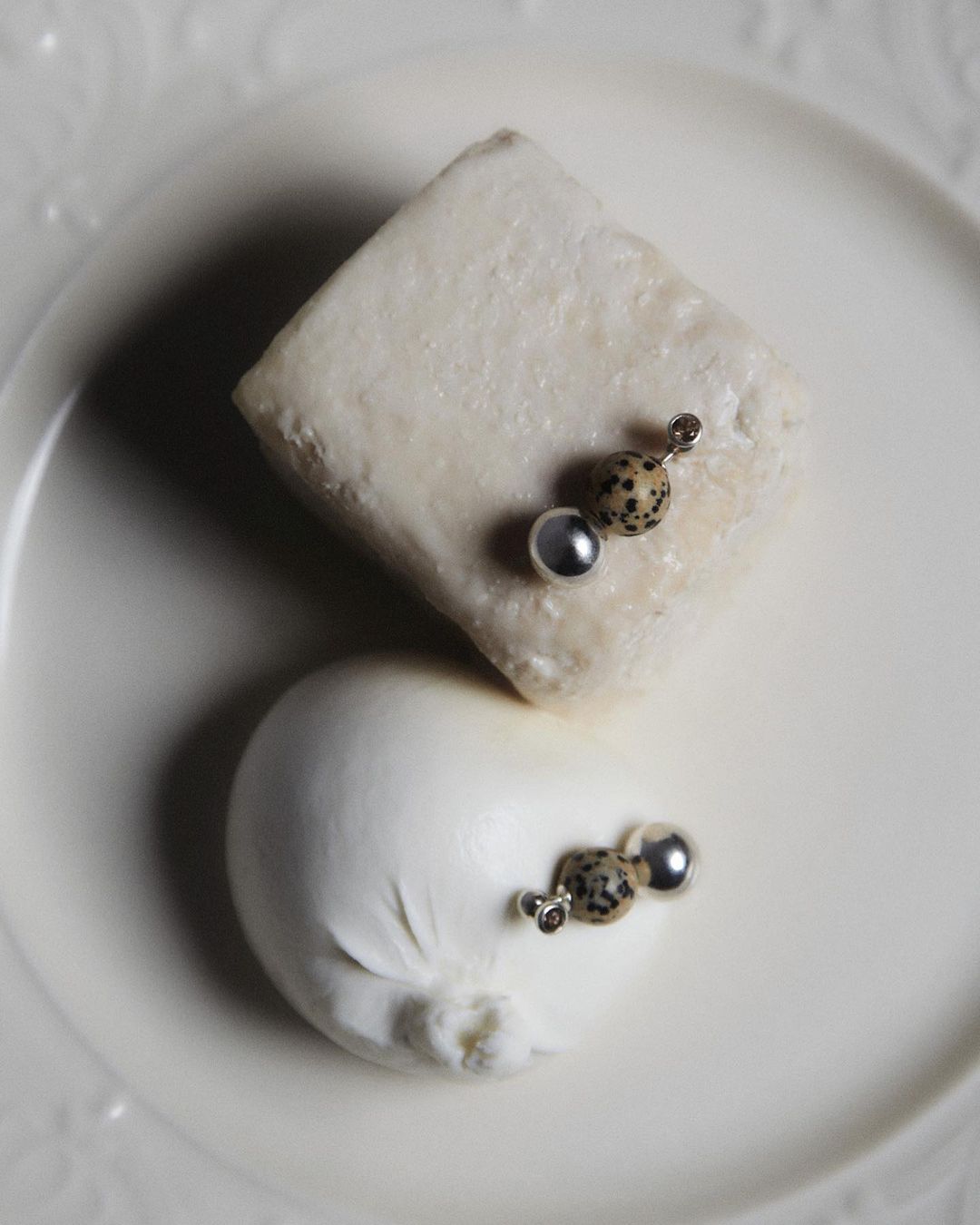Overhead shot of two cheeses adorned with decorative earrings on a white plate. One cheese is square, the other round, casting soft shadows.