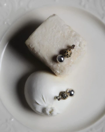 Overhead shot of two cheeses adorned with decorative earrings on a white plate. One cheese is square, the other round, casting soft shadows.