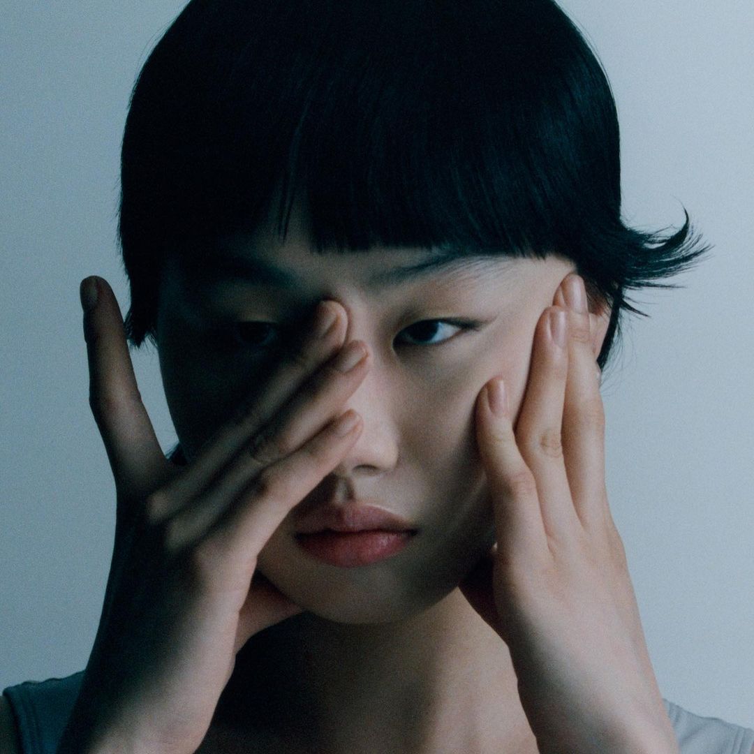 Close-up portrait of a woman with dark hair holding her hands to her face against a neutral background.