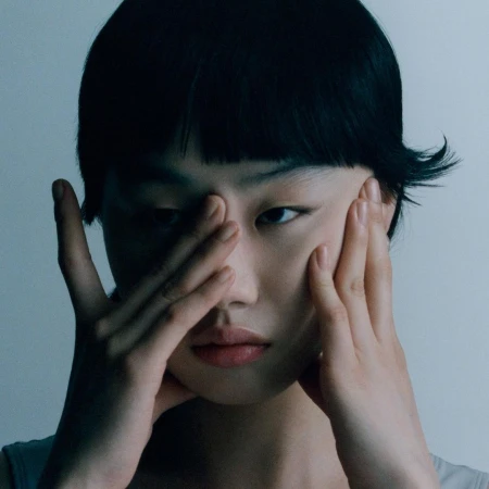 Close-up portrait of a woman with dark hair holding her hands to her face against a neutral background.