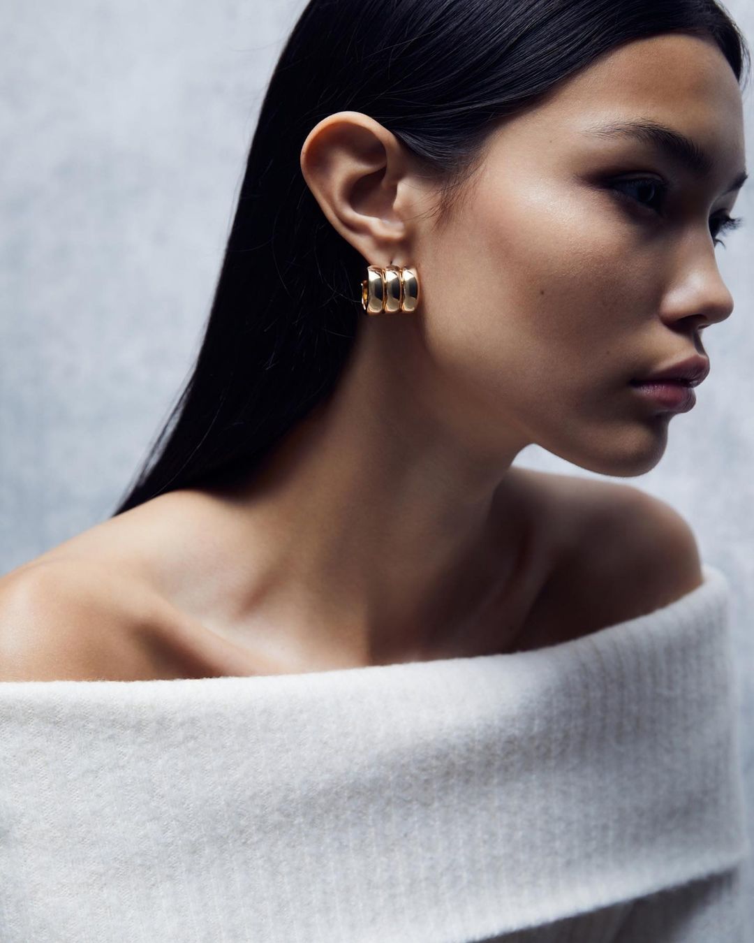 Close-up shot of an adult female wearing a white off-the-shoulder sweater and golden hoop earrings against a grey background.
