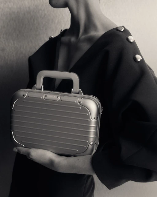 A sleek, silver metallic mini suitcase is held by a person in a black dress, emphasizing a blend of fashion and travel accessory.