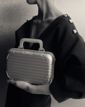 A sleek, silver metallic mini suitcase is held by a person in a black dress, emphasizing a blend of fashion and travel accessory.