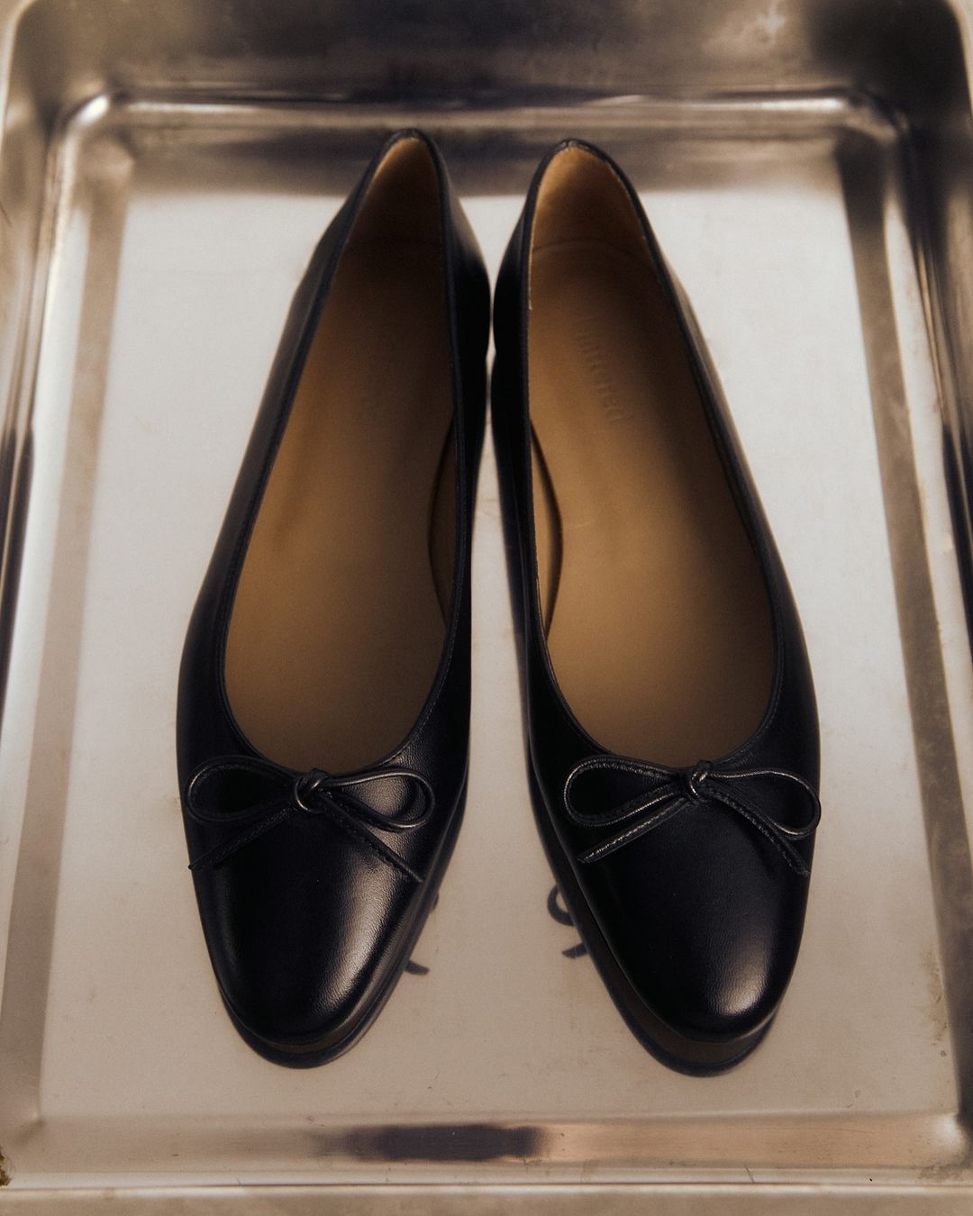 Close-up of two black leather ballet flat shoes with small bows, arranged on a silver tray. Simple and elegant footwear.
