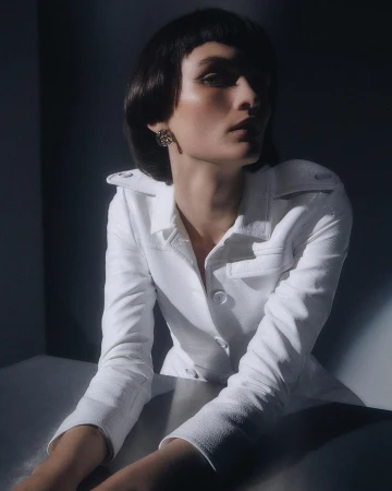 Model with dark short bangs wearing a white collared jacket leaning on a table in low-key lighting by Daisy Walker