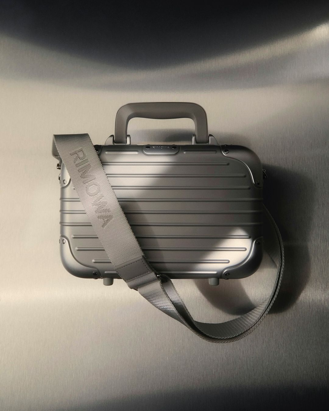Silver Rimowa Personal Crossbody Aluminum Case with strap, small luggage with handle for travel, against a neutral background.