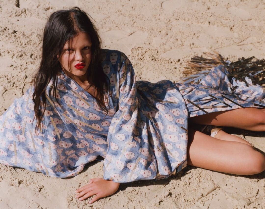 A woman in a patterned robe lies on tan sand with bright red lipstick, dark hair, and a relaxed pose in the sandy outdoor setting.