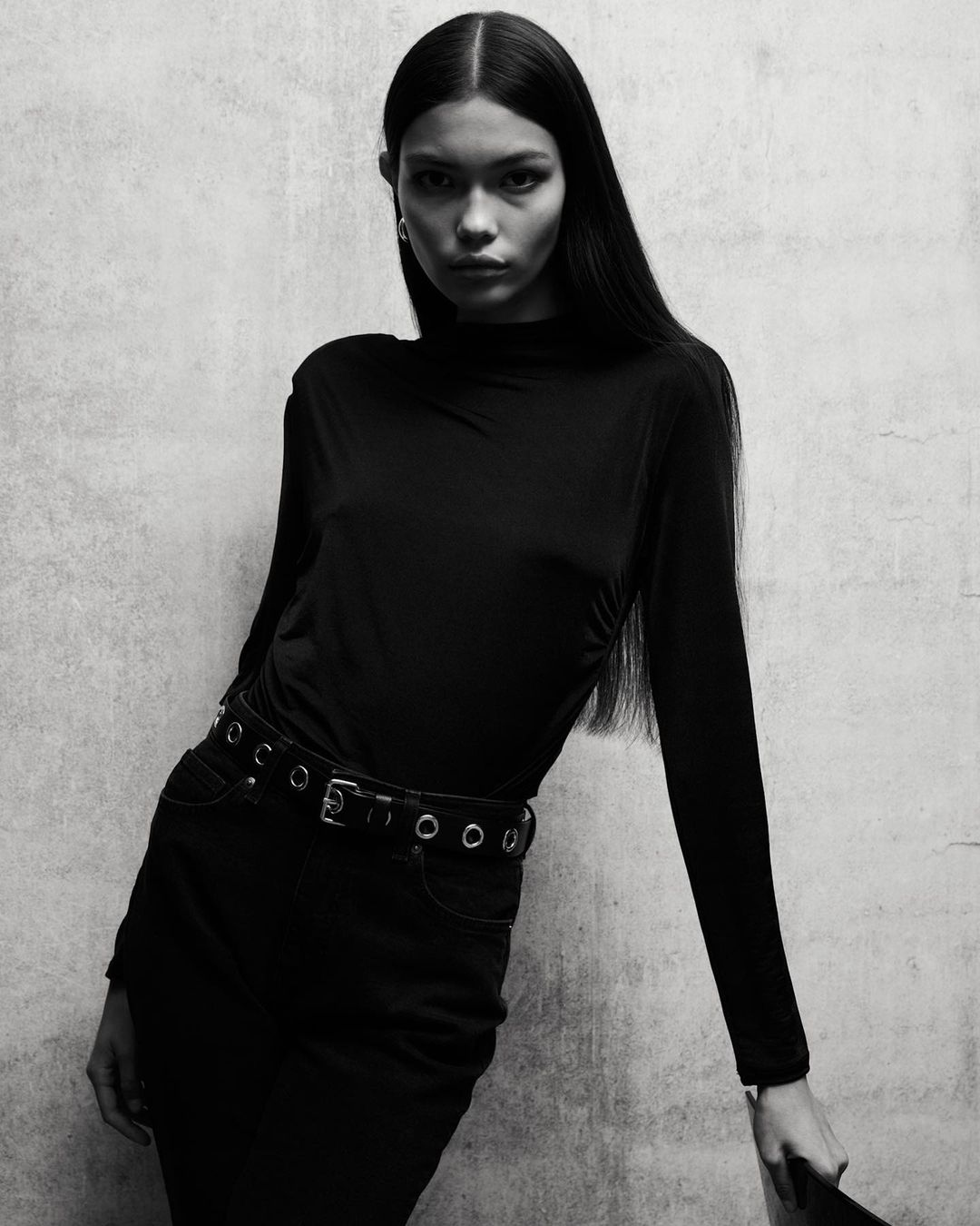 Monochrome portrait of a young woman in a long-sleeved top and skirt, leaning against a textured wall, looking directly at the camera.