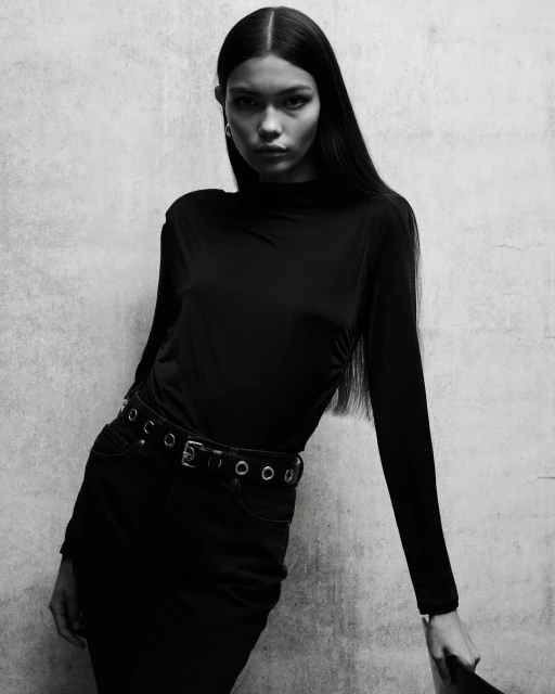 Monochrome portrait of a young woman in a long-sleeved top and skirt, leaning against a textured wall, looking directly at the camera.