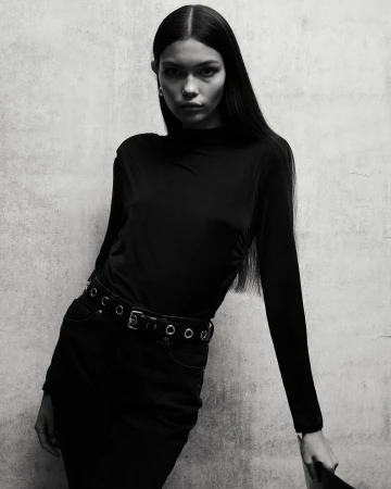 Monochrome portrait of a young woman in a long-sleeved top and skirt, leaning against a textured wall, looking directly at the camera.