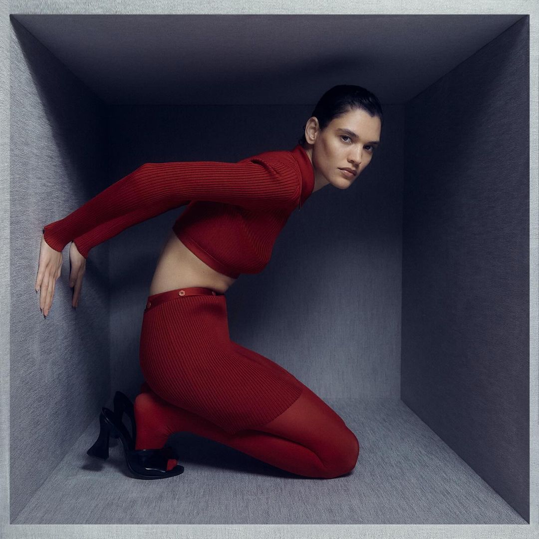Model in red turtleneck and red trousers kneeling inside a grey cubic set by Daisy Walker