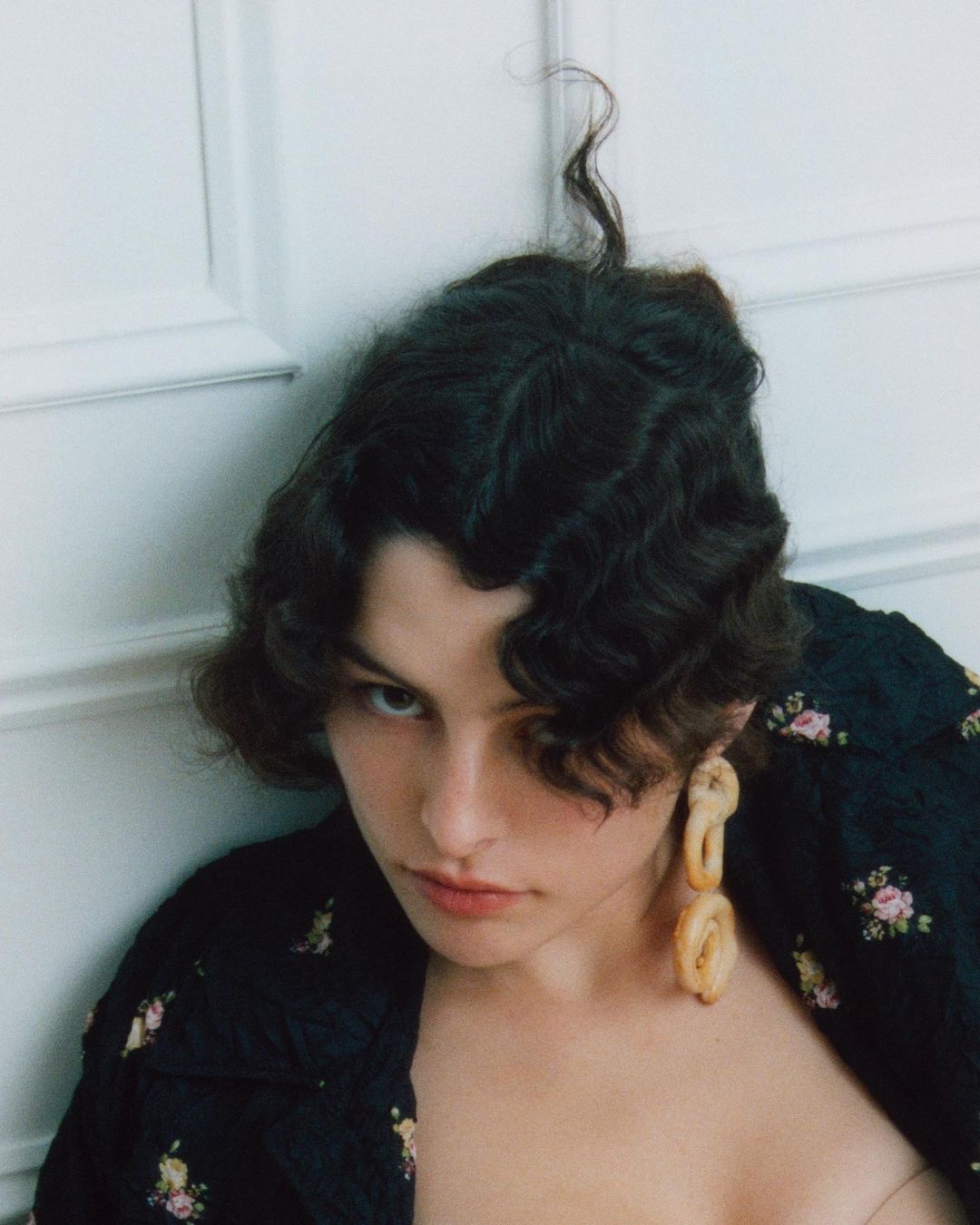 Portrait of a young adult female with brown wavy hair, wearing a black floral jacket and unique sculptural earrings, indoors against a white wall.