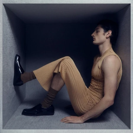 Man wearing a ribbed beige sleeveless top and flared trousers seated inside a grey cube structure