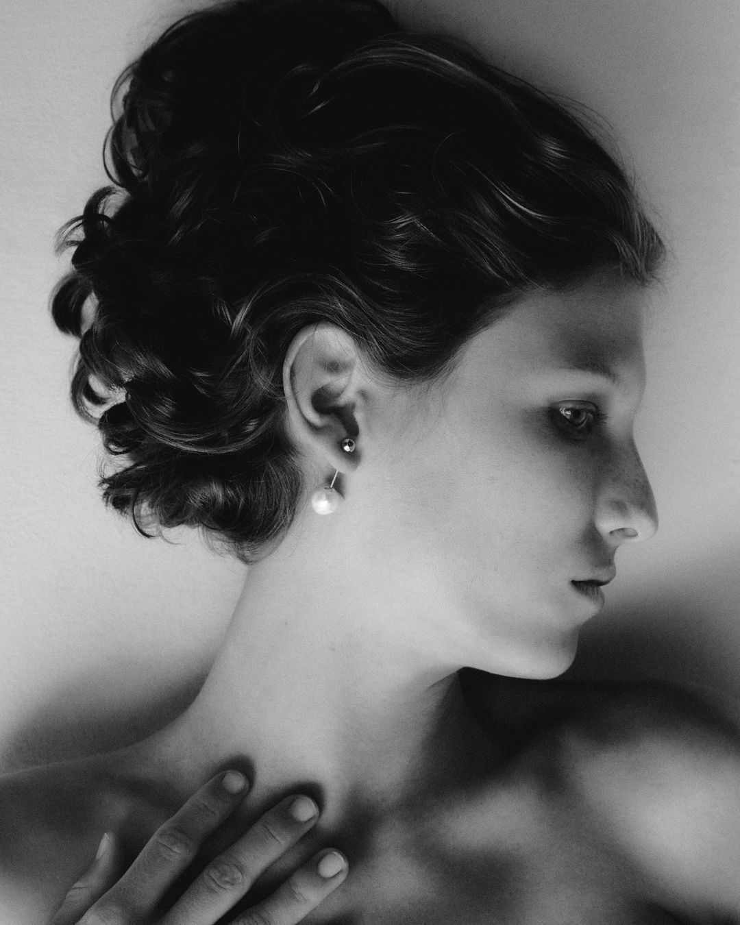 Black and white portrait of a woman with dark hair in an updo, side profile, wearing pearl earrings, hand resting on chest, eyes looking off-frame.