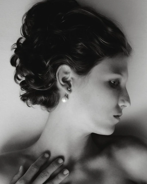 Black and white portrait of a woman with dark hair in an updo, side profile, wearing pearl earrings, hand resting on chest, eyes looking off-frame.