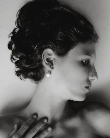 Black and white portrait of a woman with dark hair in an updo, side profile, wearing pearl earrings, hand resting on chest, eyes looking off-frame.