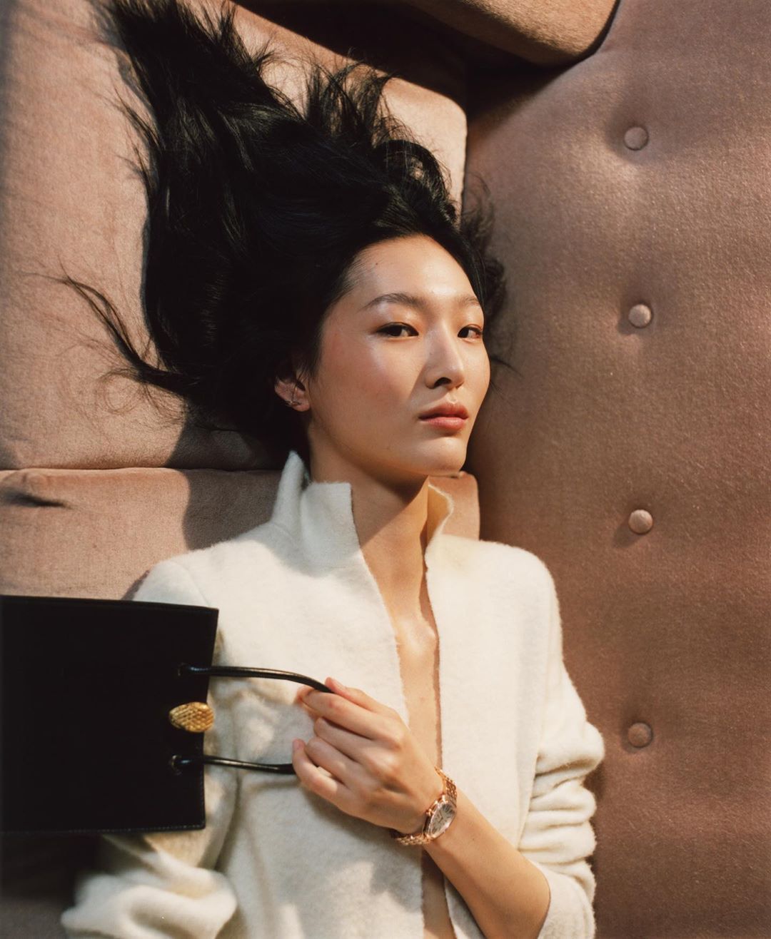 Woman with dark hair lies on tan couch wearing white sweater, a gold wristwatch, and holding a small black purse with gold buttons.